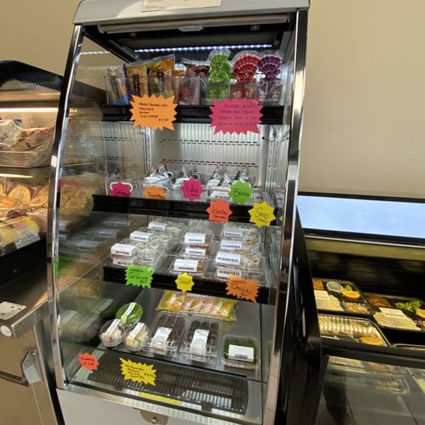 A refrigerated display case at FuruSato Bento filled with various Japanese snacks, sushi, and desserts.