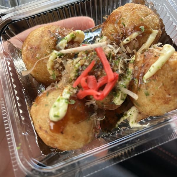 Four takoyaki, drizzled with sauce and mayonnaise, topped with red ginger and bonito flakes in a plastic container.